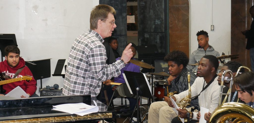 A man, standing, talks to a group of young seated students holding musical instruments