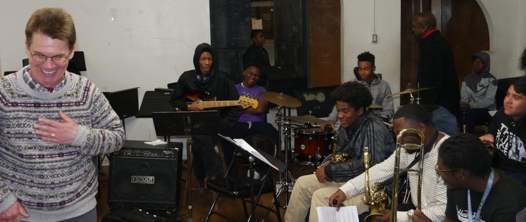 A man, standing, and a group of seated students holding musical instruments all laugh.