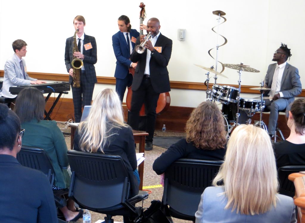 Five jazz musicians hold instruments and perform for a group of seated people