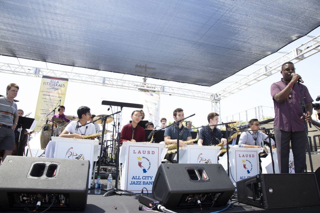 A man in a purple shirt stands with a microphone before a group of seated high school students with musical instruments