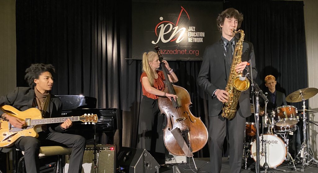 Four students playing jazz instruments perform on a stage with a black backdrop.