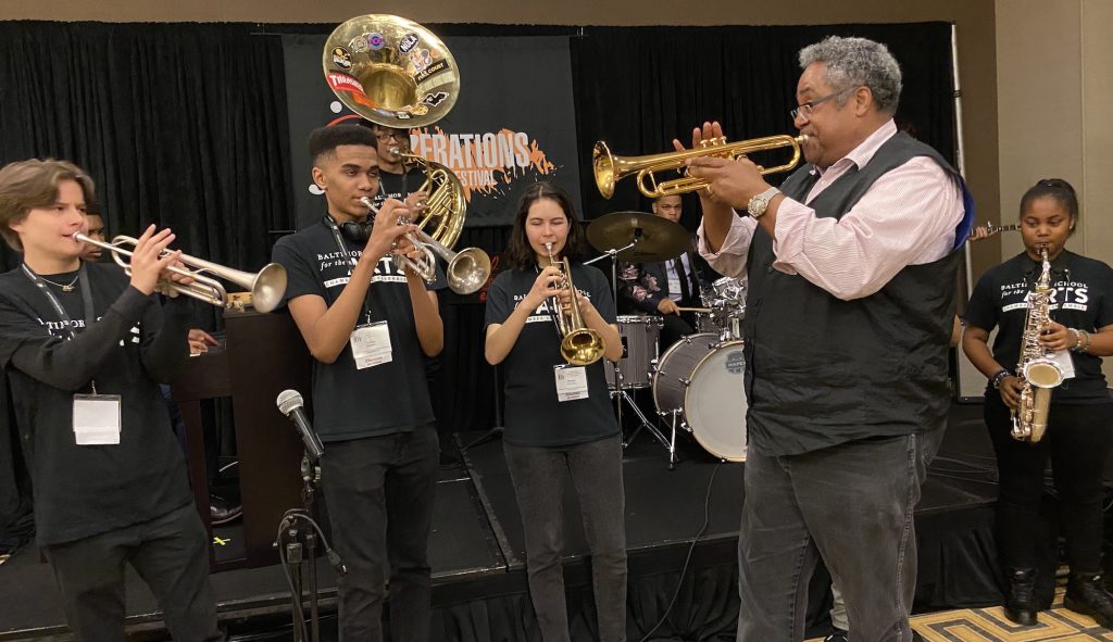 A man stands in the foreground playing his trumpet with a group of students, also playing instruments.