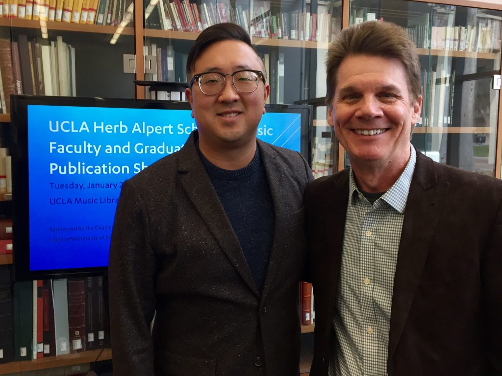 Two men stand smiling in a library in front of a television monitor